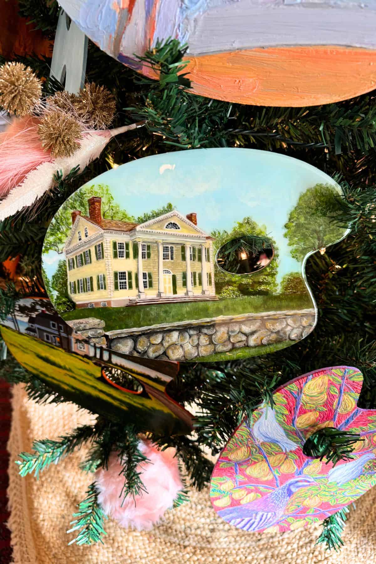 A decorated Christmas tree features artist palettes, one painted with a yellow colonial-style house and a stone wall, displayed among ornaments and greenery.