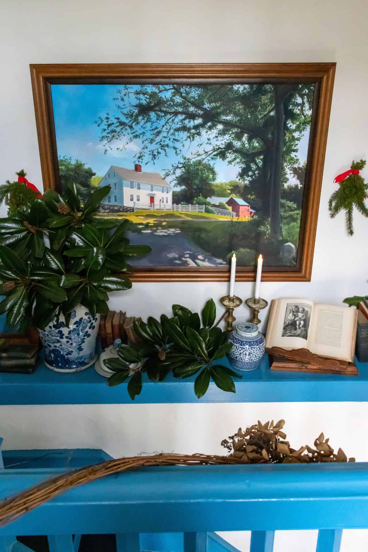 A framed painting of a house hangs above a shelf with potted plants, books, candles, an open book, and two small wreaths on the wall.