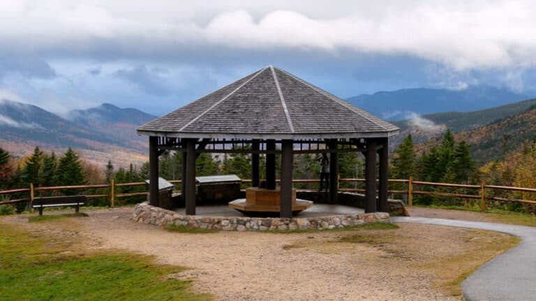 A wooden gazebo with a pyramid-shaped roof stands on a stone base, overlooking a scenic mountain landscape with autumn foliage and a cloudy sky.