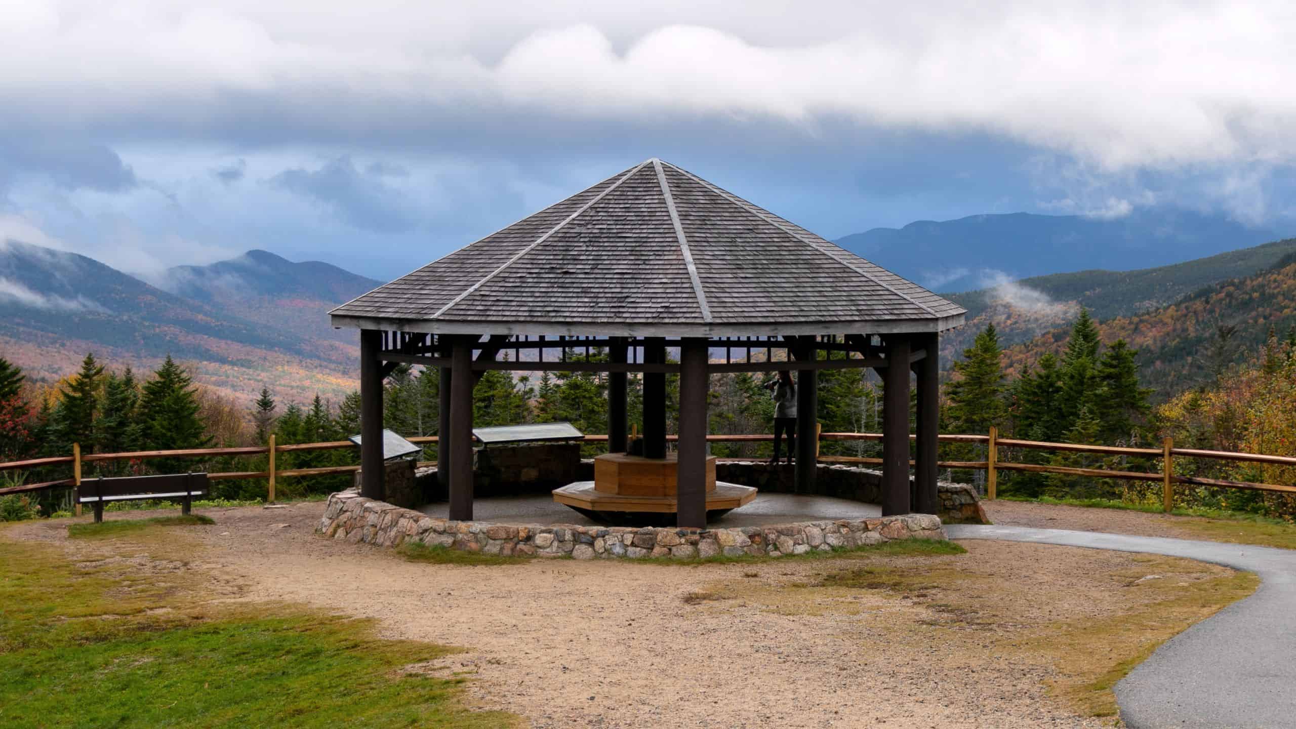 A wooden gazebo with a pyramid-shaped roof stands on a stone base, overlooking a scenic mountain landscape with autumn foliage and a cloudy sky.