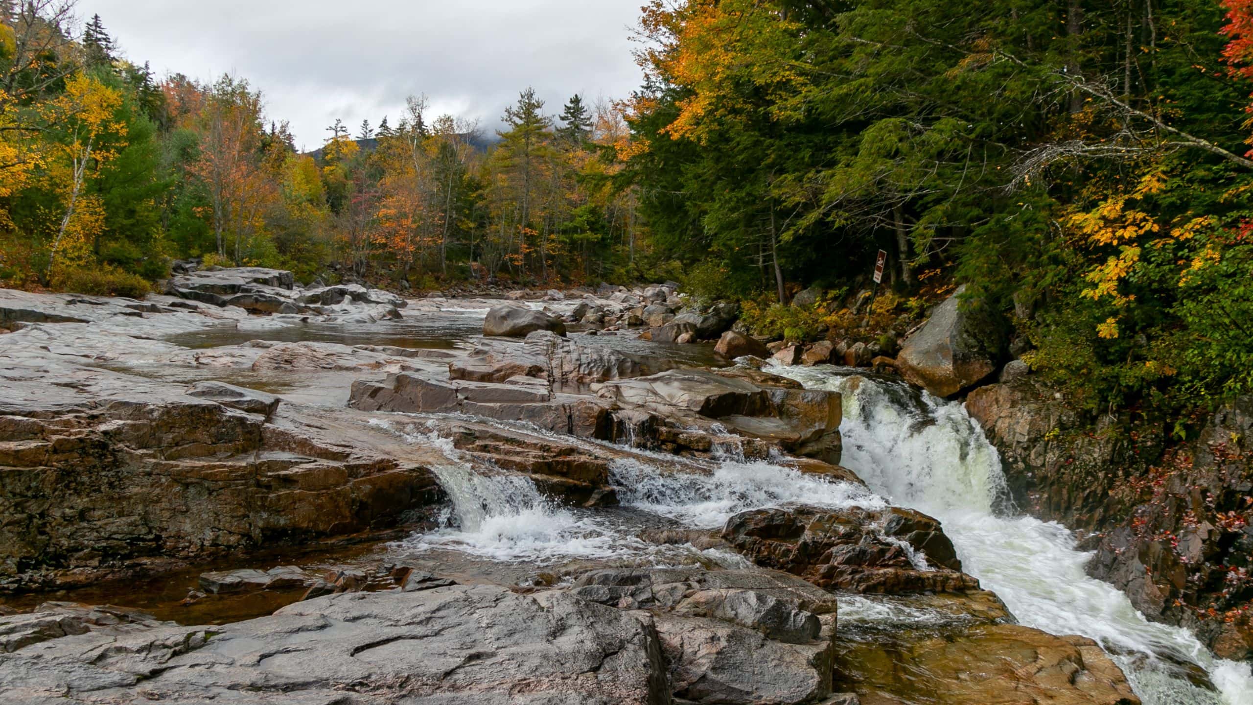 A small waterfall flows over rocky terrain surrounded by trees with autumn foliage under a cloudy sky.