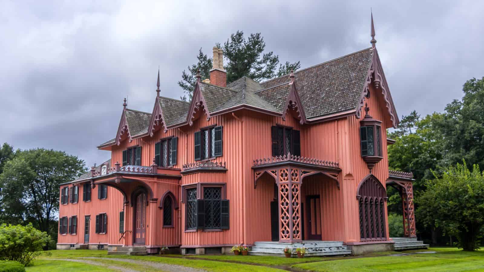A large, pink Victorian-style house with ornate wooden trim, pointed gables, and a steep roof sits on a green lawn under a cloudy sky.