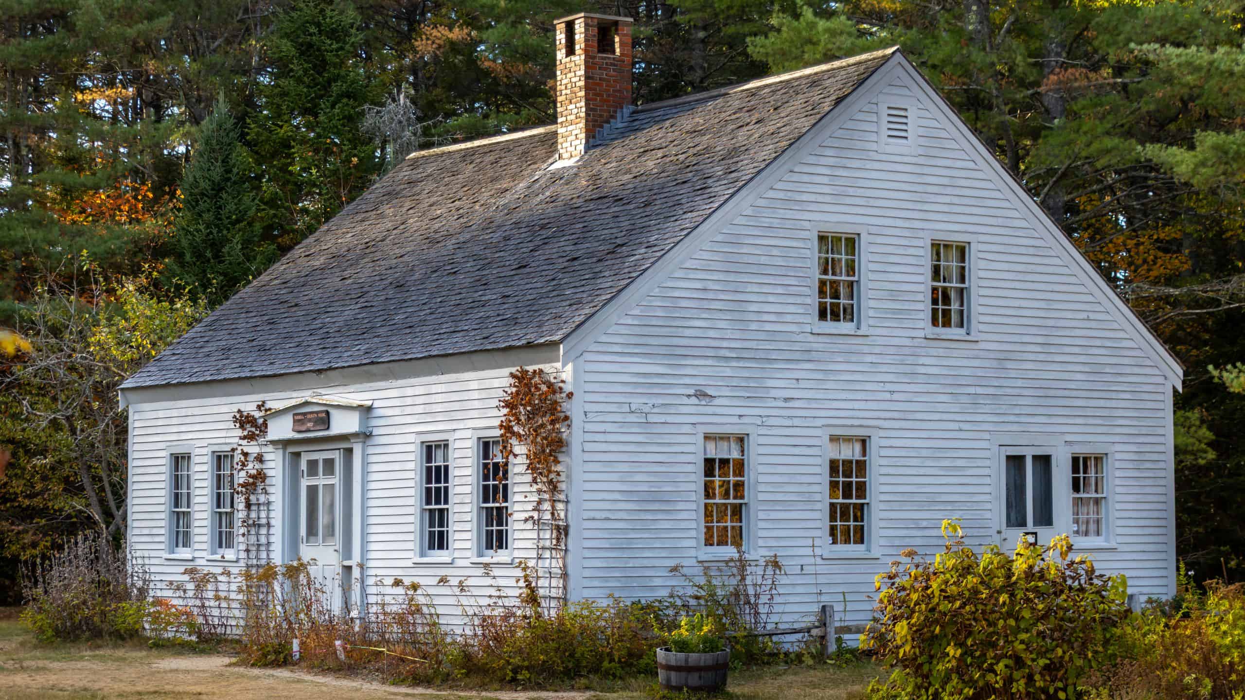A small, white wooden house with a steep roof, chimney, and multiple windows, surrounded by trees and overgrown bushes.