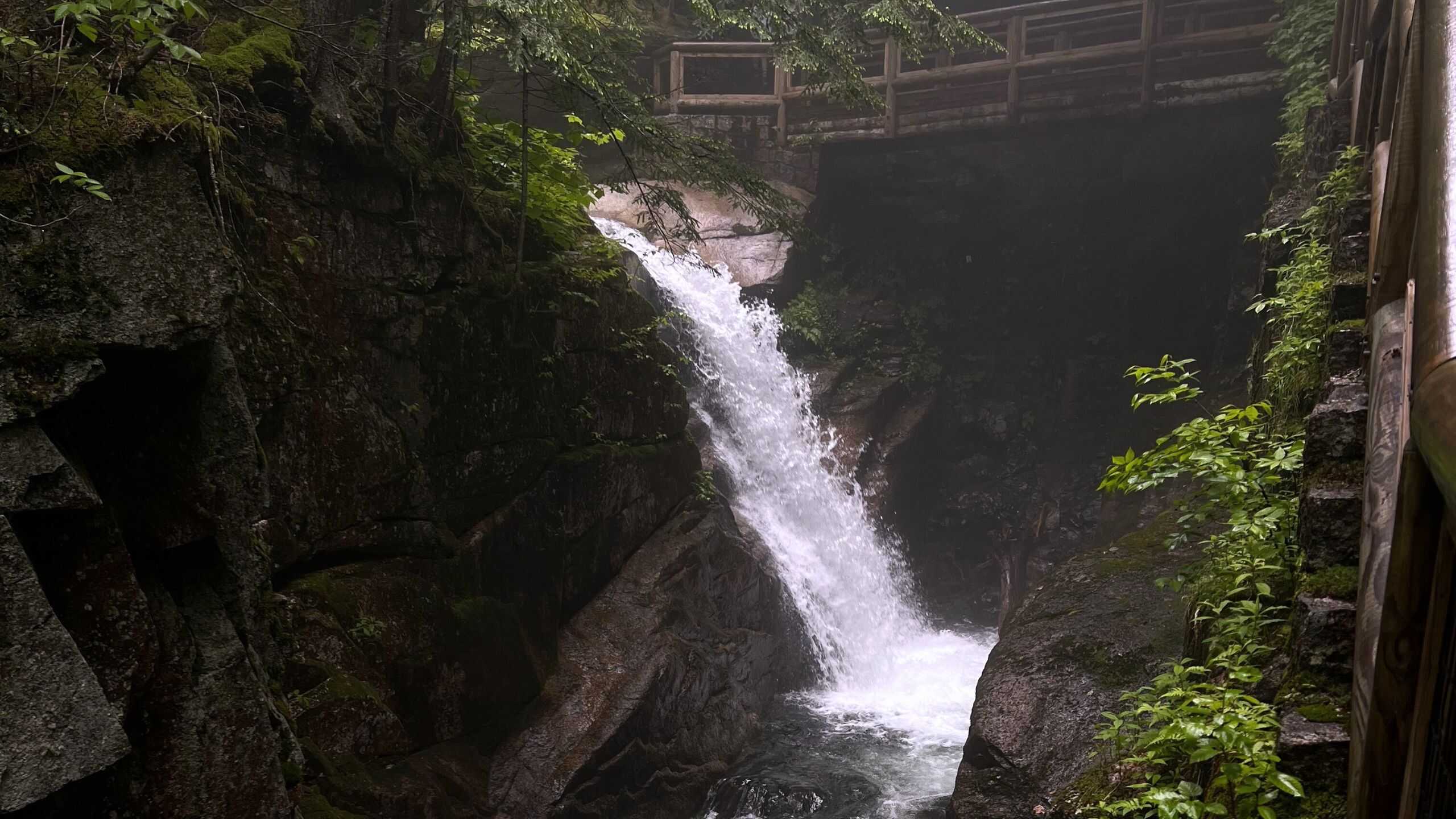 A small waterfall flows between rocky cliffs surrounded by greenery, with a wooden footbridge above and mist rising from the water.