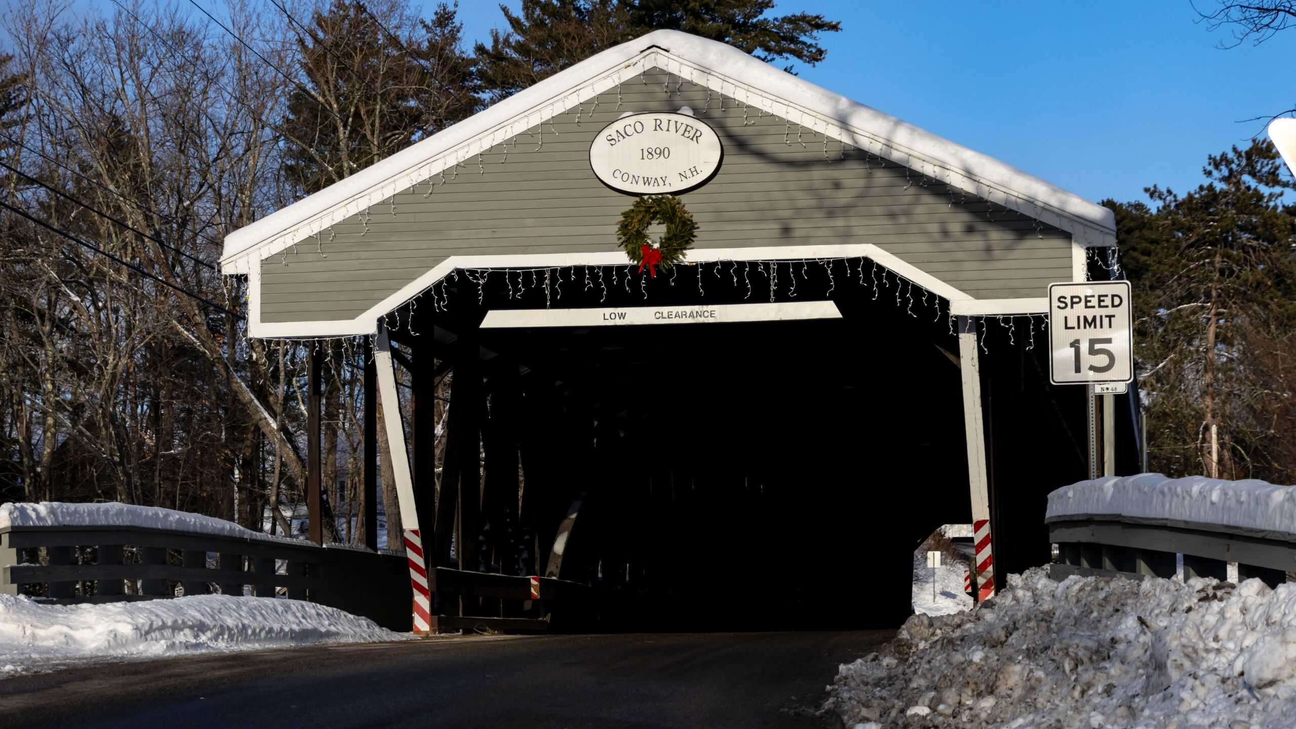 A covered wooden bridge labeled "Saco River 1890 Conway, NH" decorated with a wreath, surrounded by snow; a speed limit 15 sign is visible.