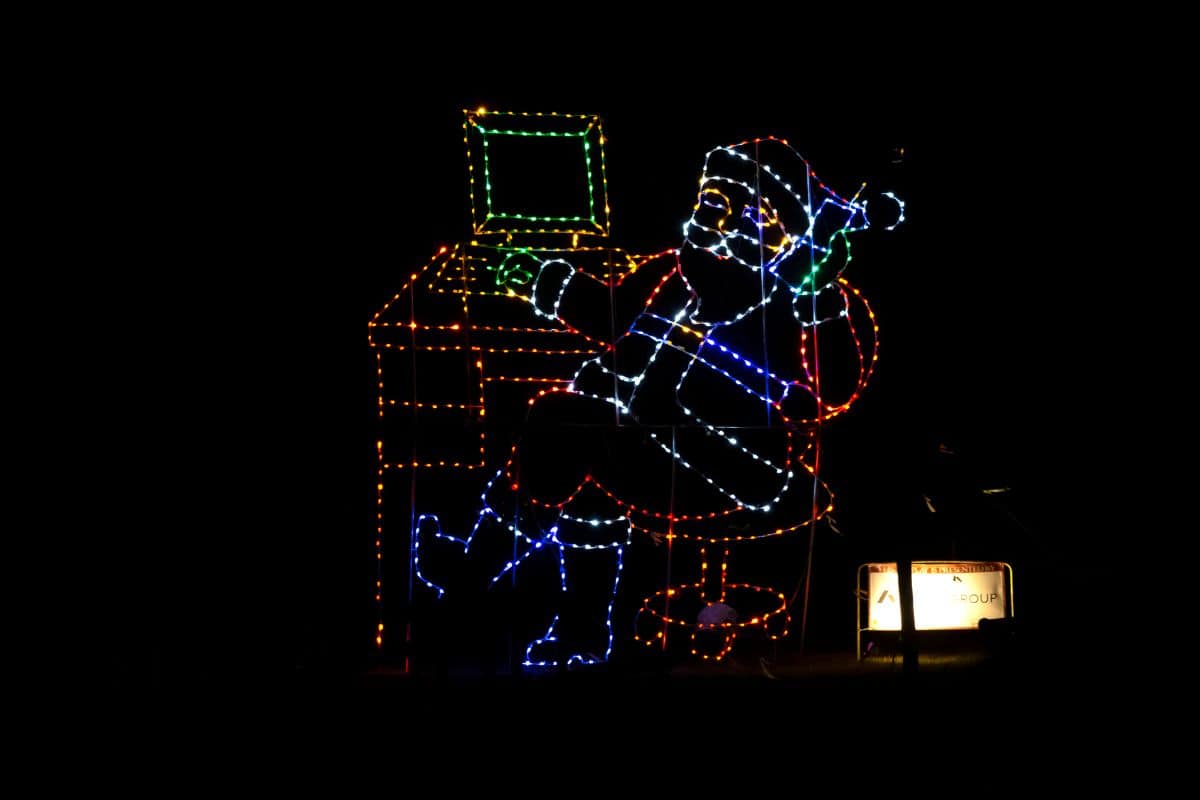 Neon light display of Santa Claus sitting at a desk, using a computer and holding a phone, against a black background.