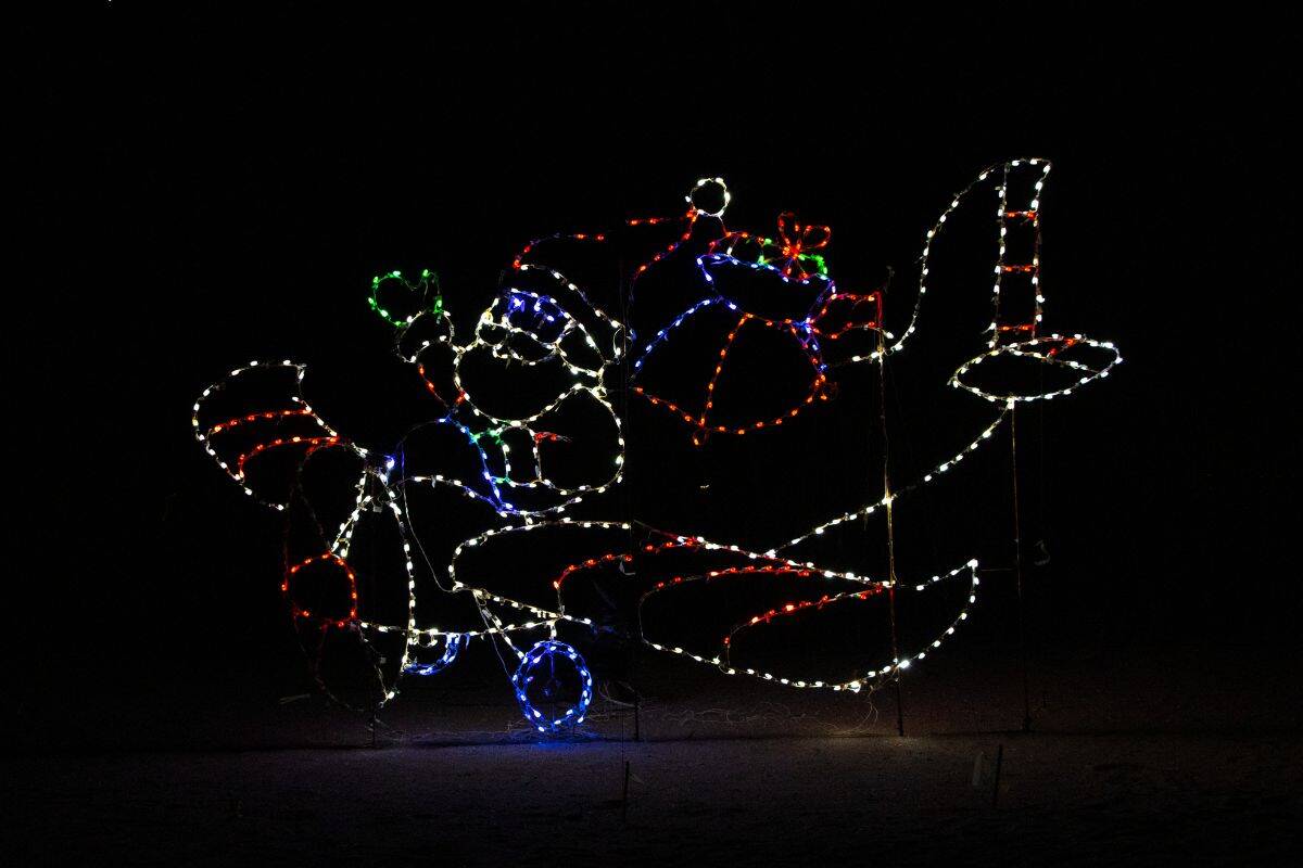 Light display showing Santa Claus in an airplane, outlined in red, white, blue, and green lights, with Santa holding a green object and a large gift behind him.