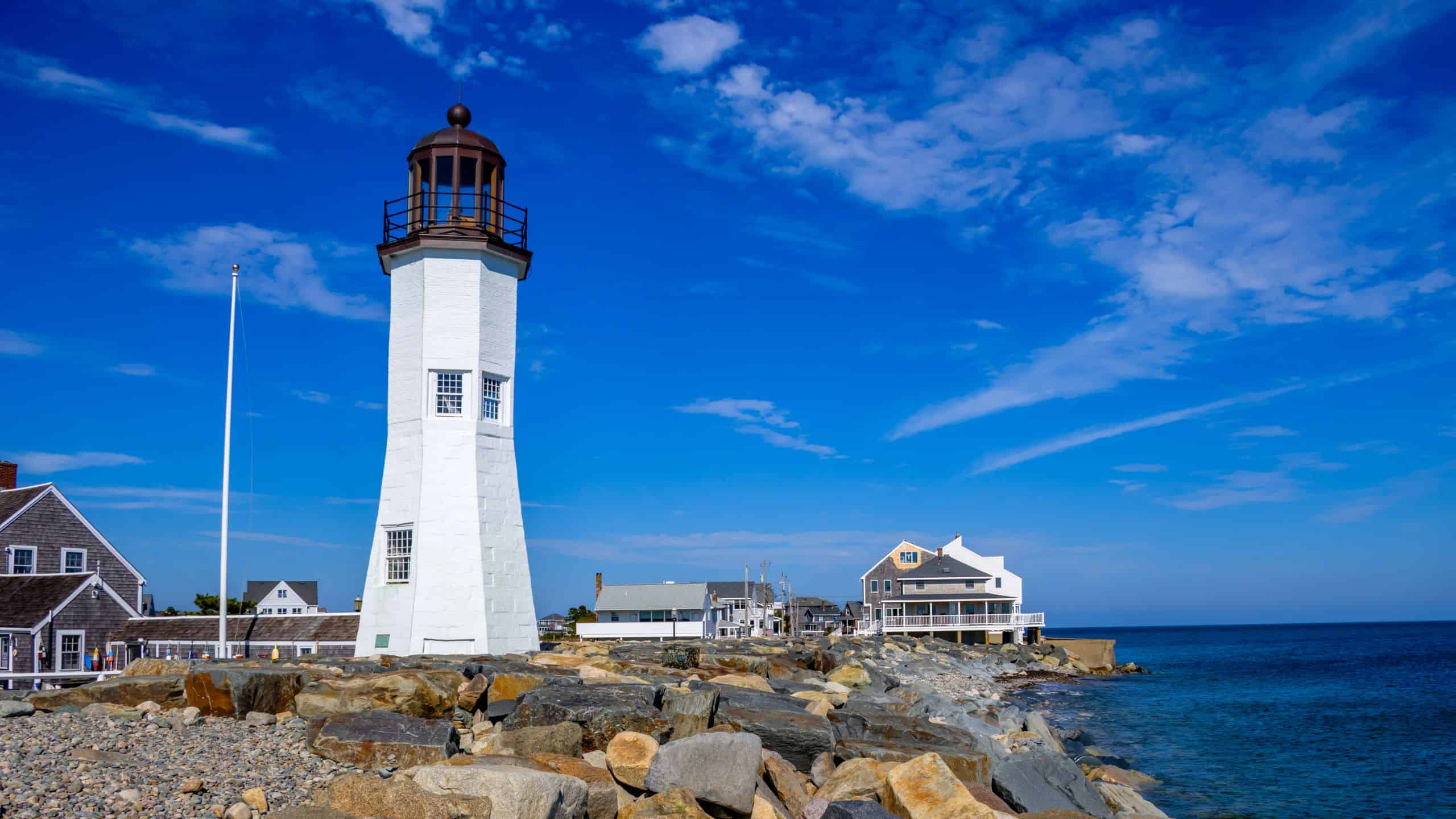 White lighthouse stands on a rocky shore with nearby coastal houses under a blue sky, overlooking the ocean—one of the iconic lighthouses in Massachusetts.