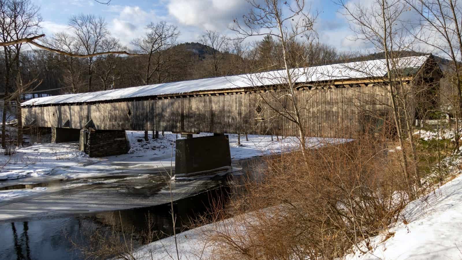 A long, wooden New England covered bridge crosses a partially frozen river in a snowy, rural landscape with leafless trees and distant hills.
