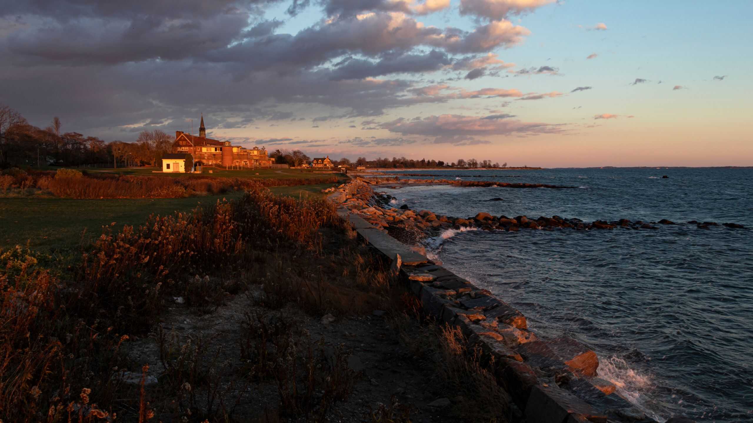 A rocky shoreline at sunset with waves hitting the rocks, grassy land on the left, and several buildings along the coast under a partly cloudy sky.
