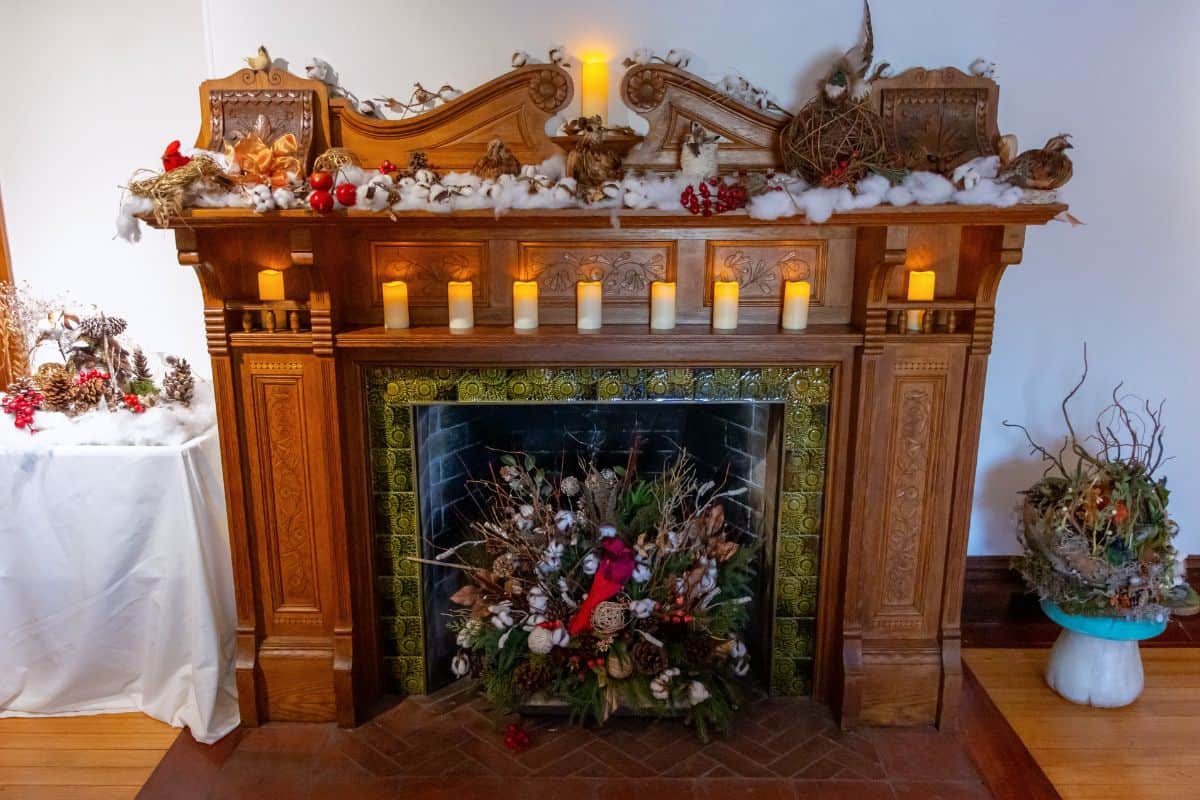 A wooden fireplace decorated with faux snow, candles, pinecones, and red accents. The hearth holds a winter-themed floral arrangement with a cardinal figure.