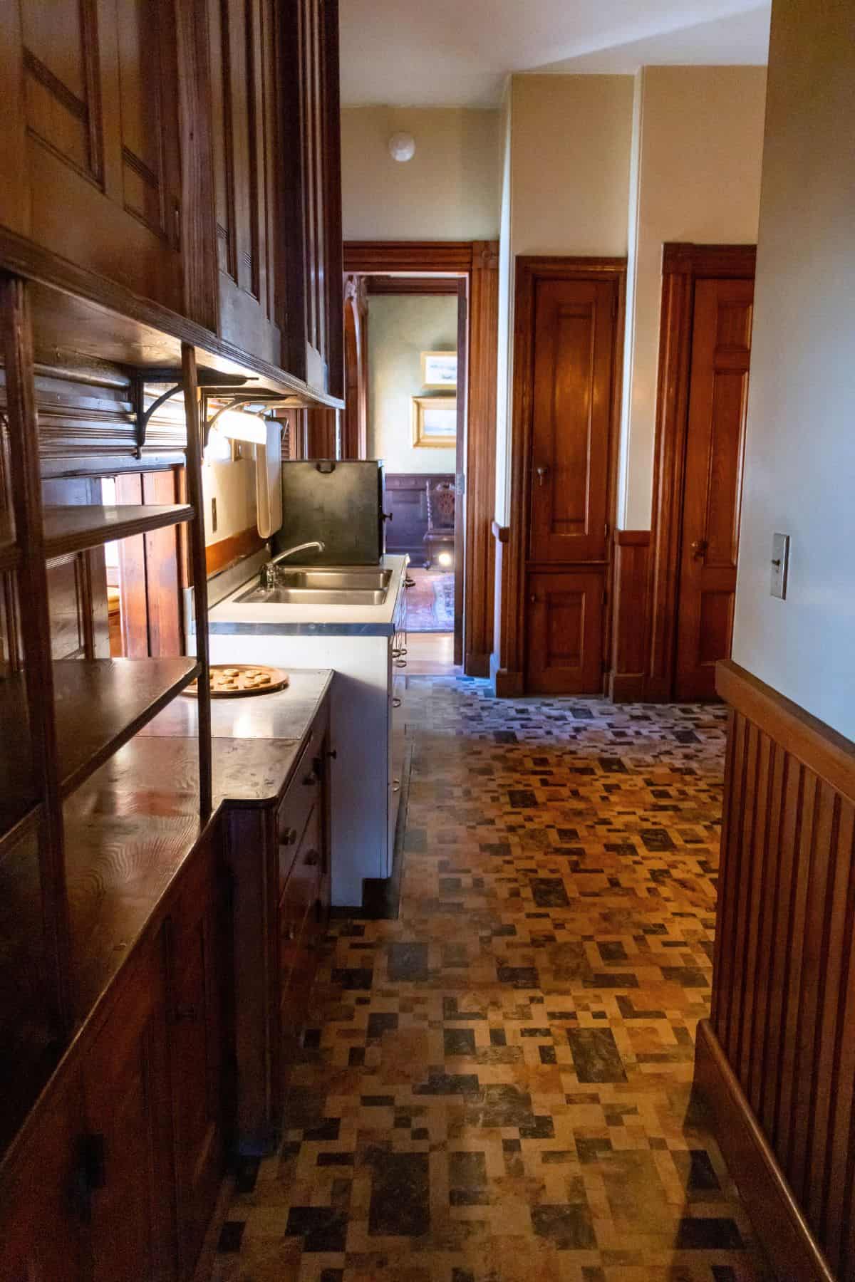 Narrow kitchen with wooden cabinets, tiled floor, a sink, and a stove, leading to a hallway with several closed wooden doors.