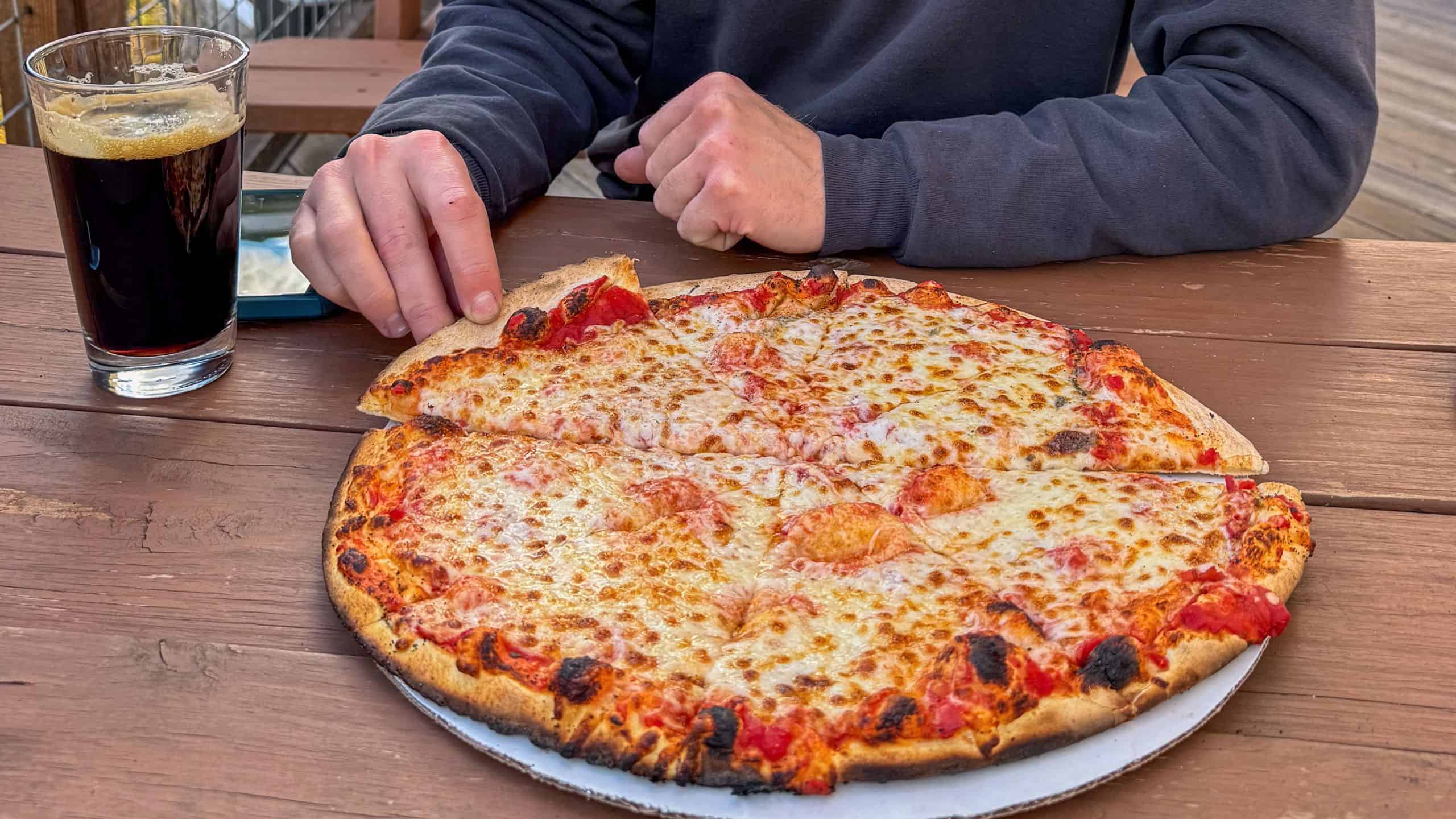 Person sitting at a wooden table with a glass of dark soda and a whole cheese pizza, about to take a slice.