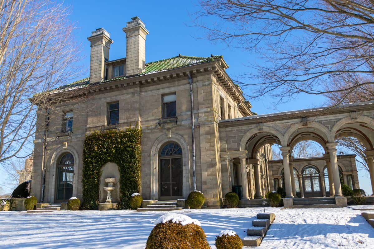 A large stone mansion with arched windows and columns stands amid snow-covered ground and leafless trees under a clear blue sky.
