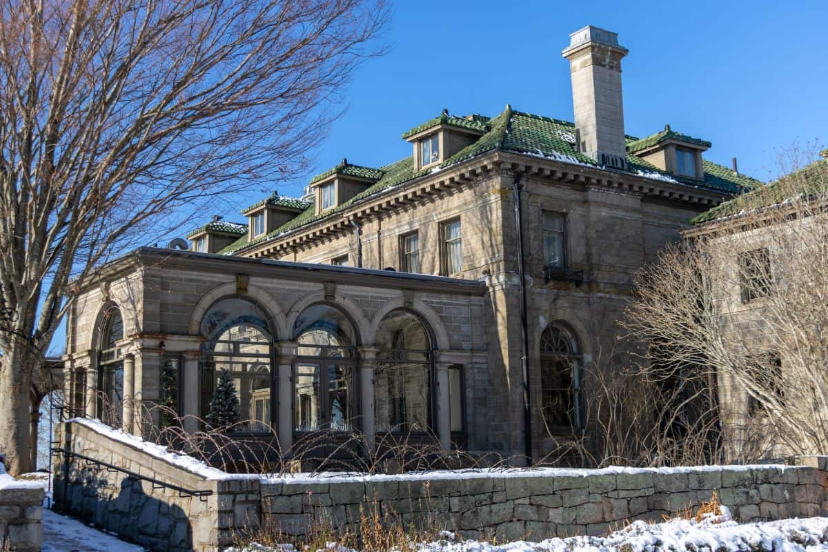 Large stone mansion with arched windows, green tile roof, and a chimney, surrounded by bare trees and patches of snow under a clear blue sky.