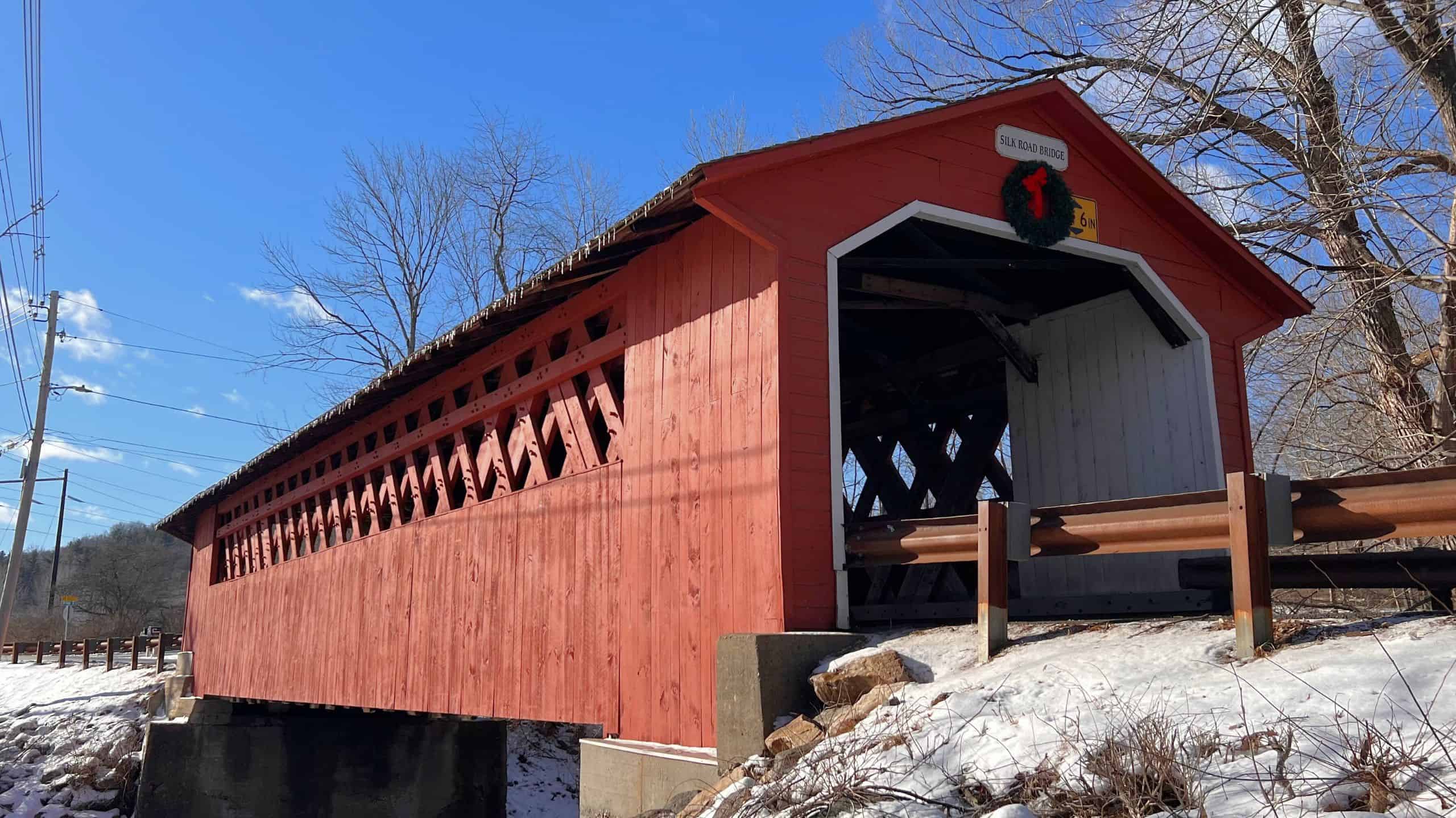 A red wooden covered bridge with lattice-style sides, reminiscent of New England covered bridges, spans a small creek surrounded by snow and bare trees under a clear blue sky.