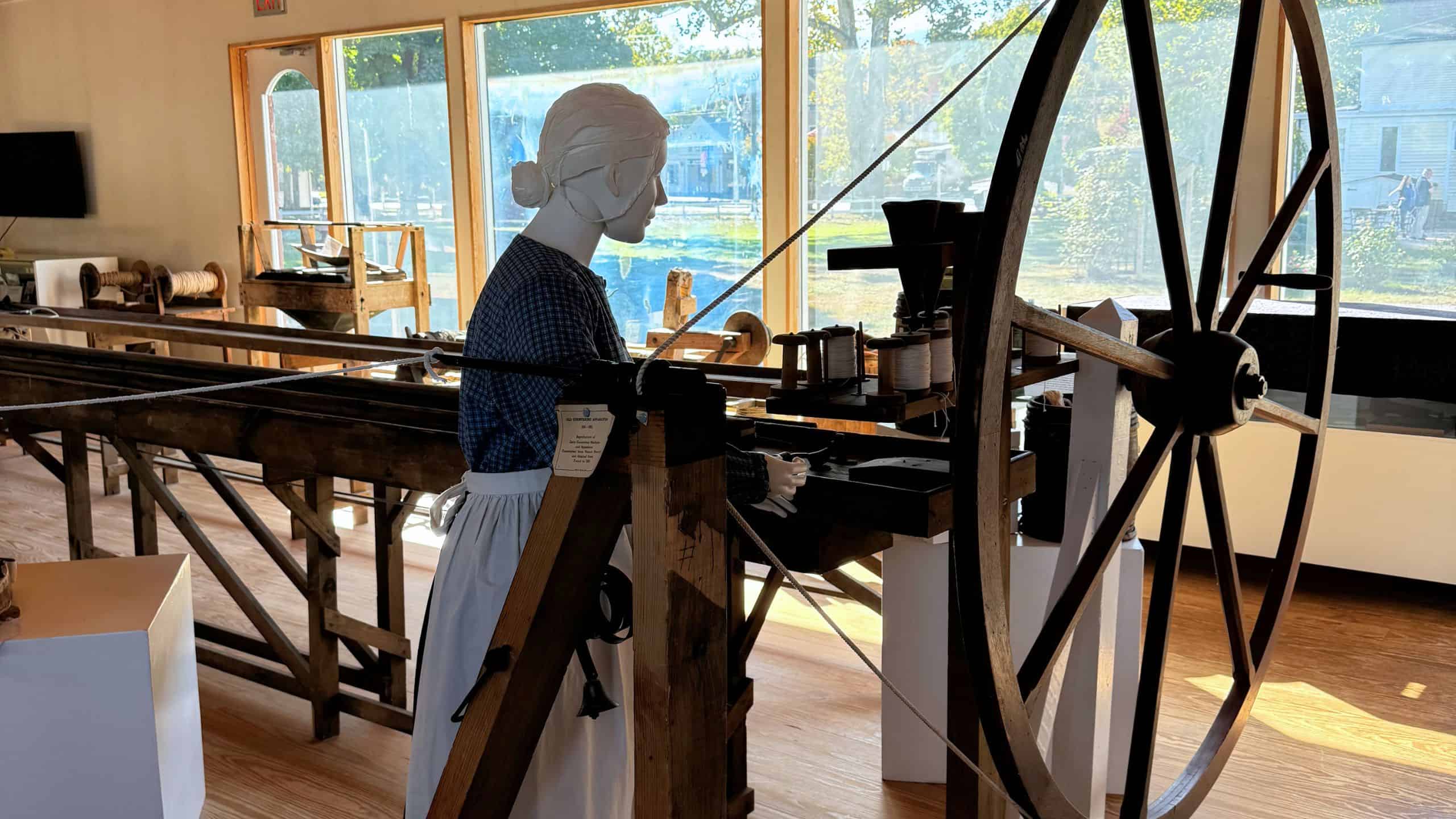 A mannequin dressed in historical clothing operates a large wooden spinning wheel in a sunlit exhibit room with large windows.