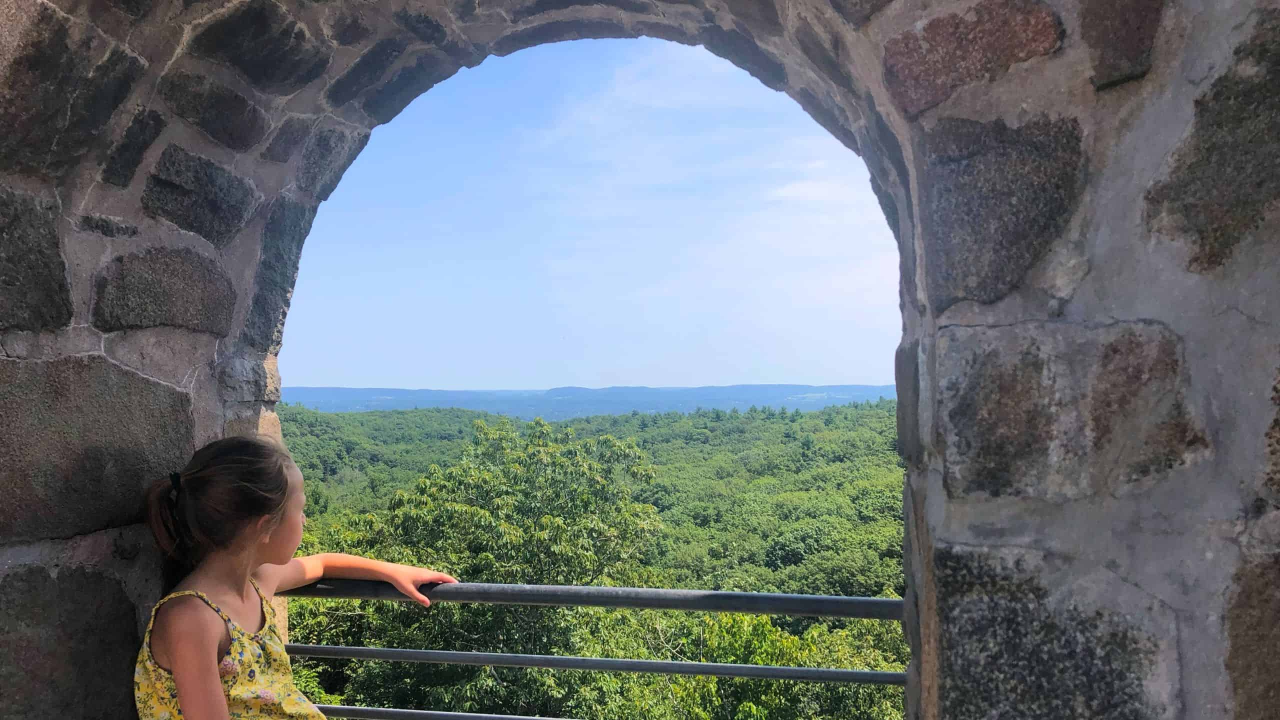 A young girl sits on a ledge inside a stone archway, looking out at a green forest landscape under a blue sky.