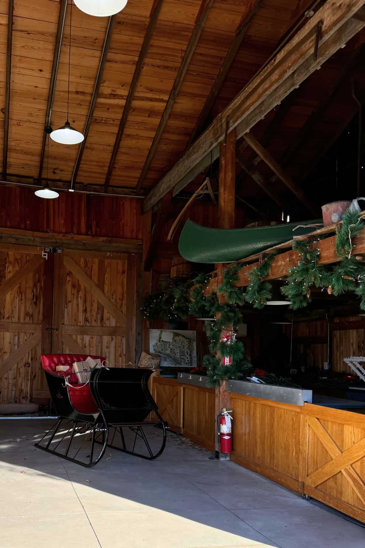 A wooden barn interior with a black and red sleigh, Christmas garland decorations, a fire extinguisher, and a canoe stored on a loft shelf.