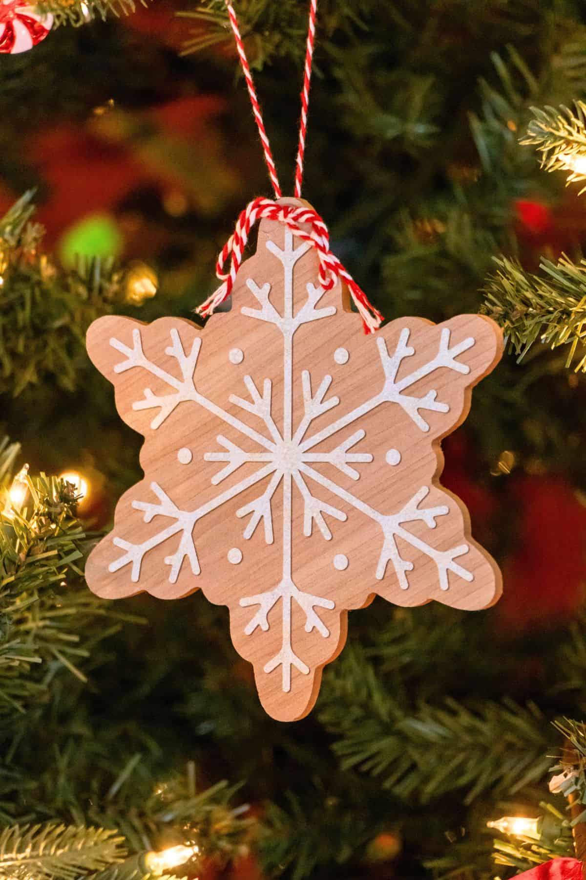 A wooden snowflake ornament with white painted details hangs on a Christmas tree by a red and white string.