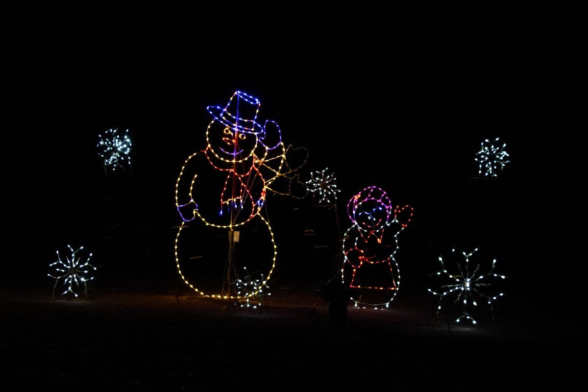 Holiday light display featuring outlined figures of a snowman and a child, surrounded by glowing starburst decorations against a dark background.