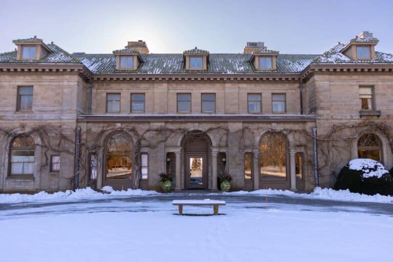 A large, historic stone mansion with arched windows and doors, surrounded by snow and bare vines, under a clear blue sky.