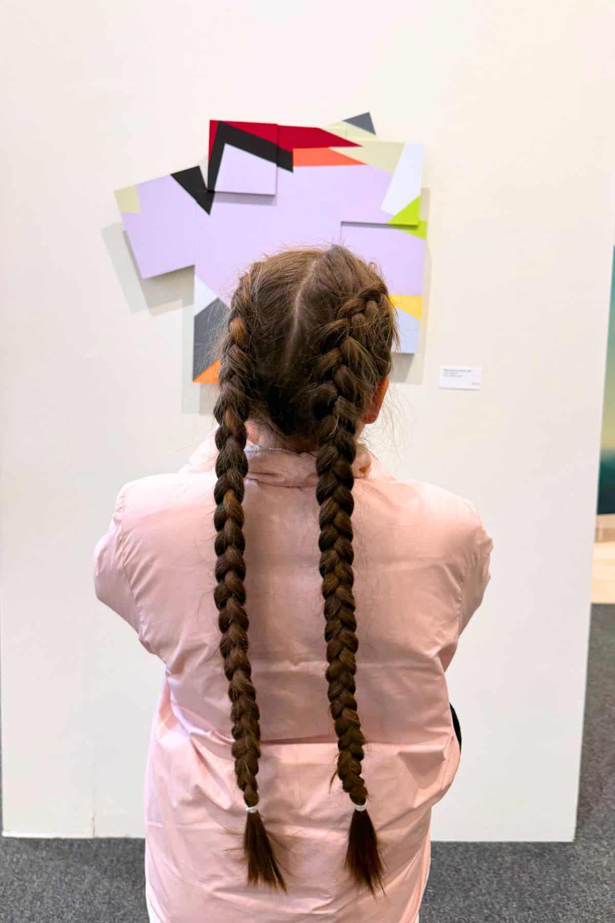 A person with two long braids, wearing a light pink shirt, stands facing an abstract geometric painting on a gallery wall.
