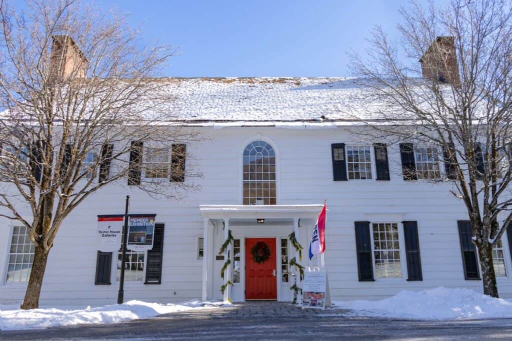 A large white building with black shutters, a red door decorated with a wreath, and snow covering the roof and ground, on a clear winter day.