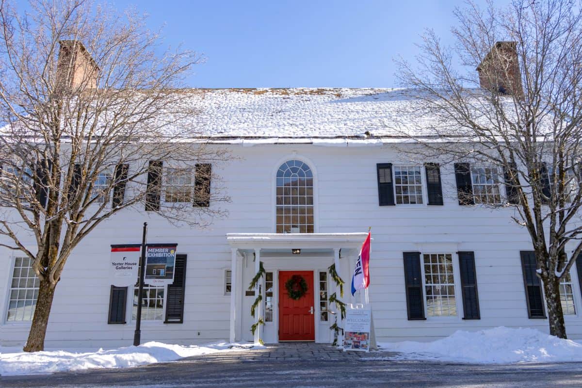 A large white building with black shutters, a red door decorated with a wreath, and snow covering the roof and ground, on a clear winter day.