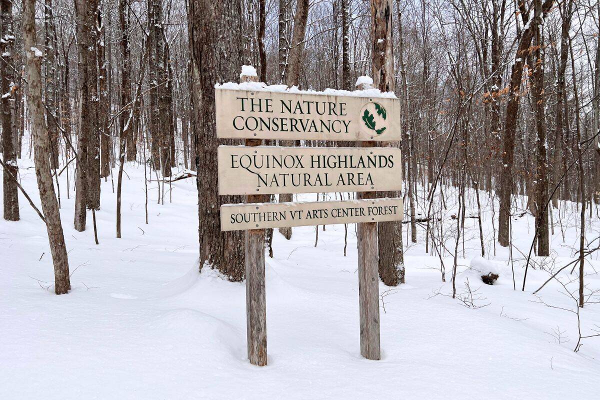 A wooden sign in a snowy forest reads: “The Nature Conservancy, Equinox Highlands Natural Area, Southern VT Arts Center Forest.”.