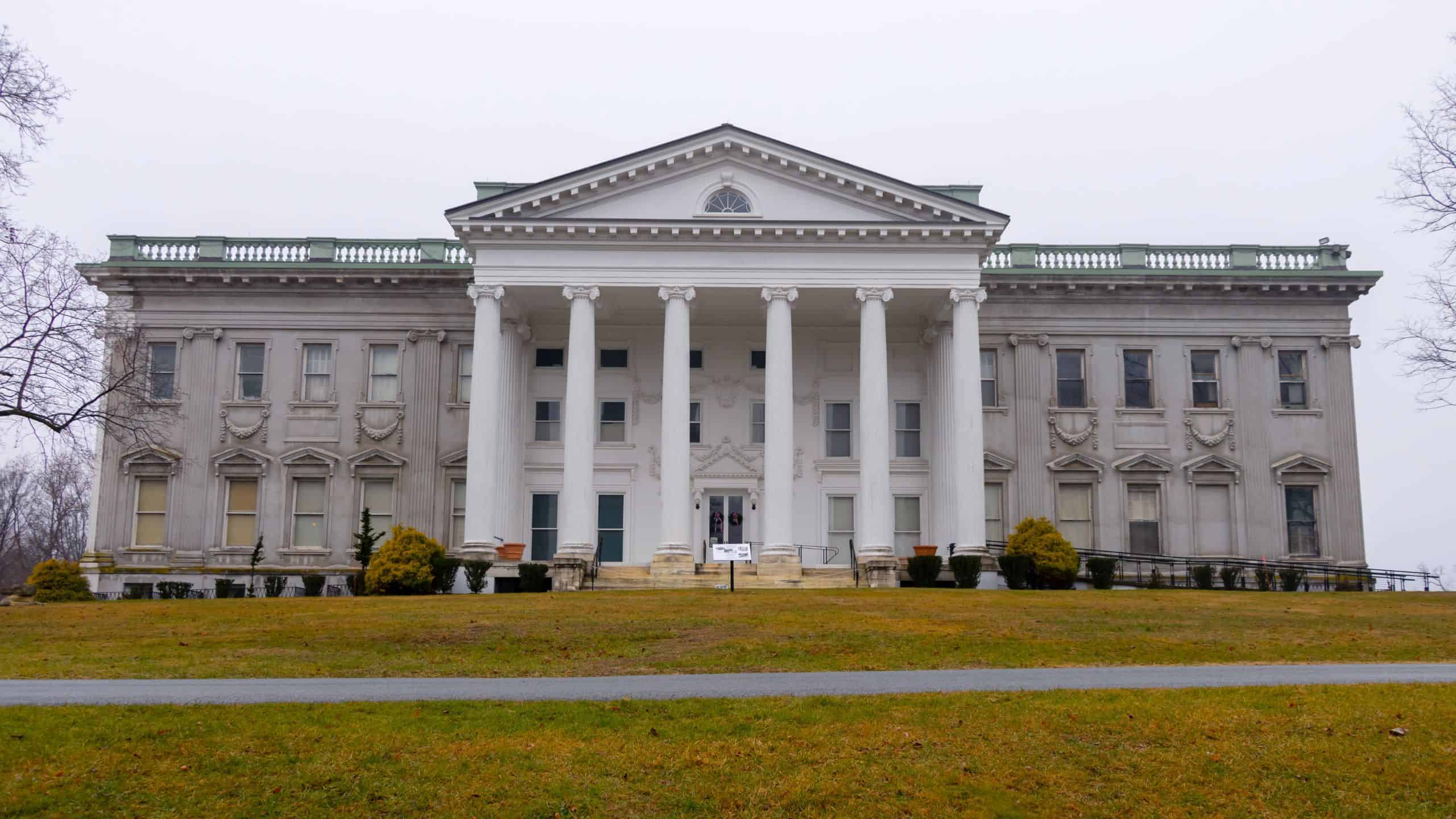 Large neoclassical building with tall white columns, symmetrical windows, and a triangular pediment, set on a grassy lawn under an overcast sky.