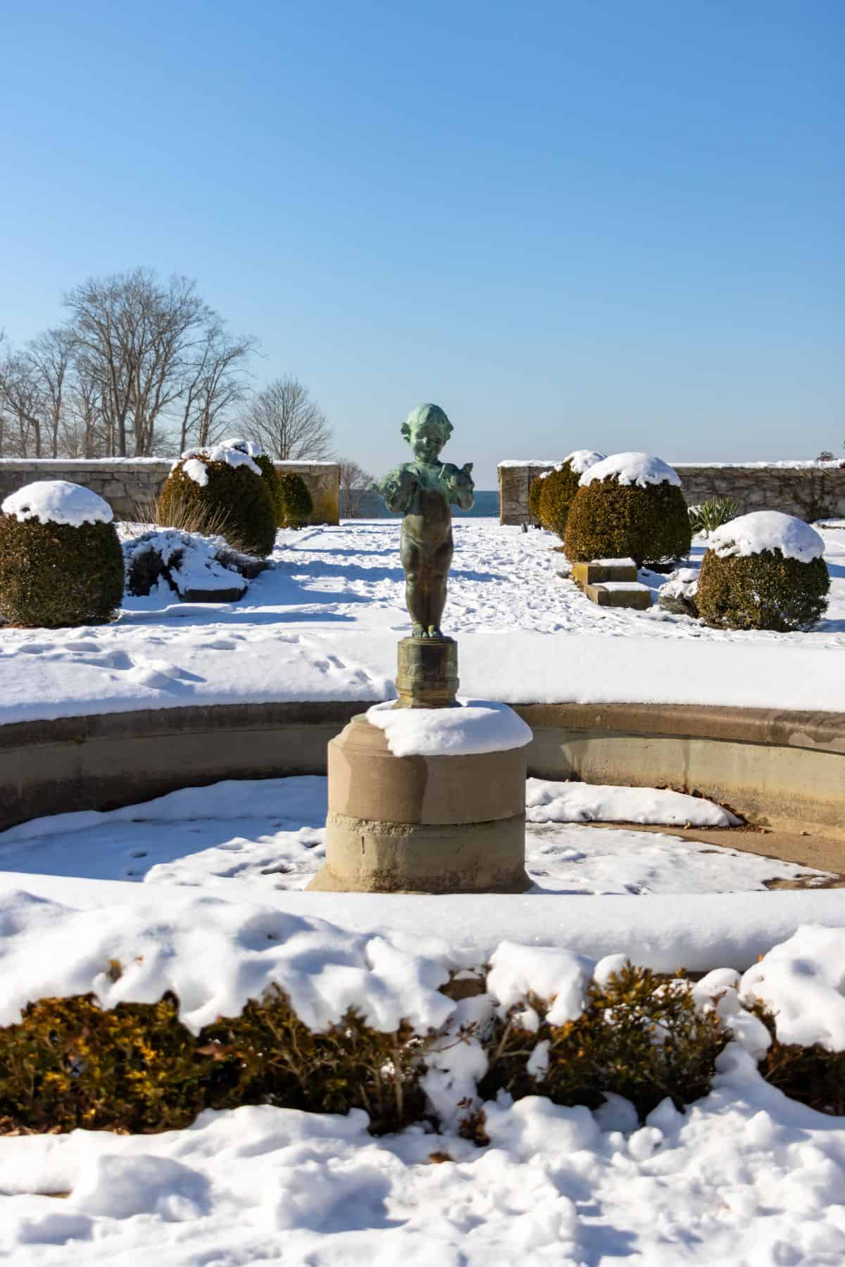 A bronze statue stands in the center of a snow-covered garden with trimmed hedges and a clear blue sky in the background.
