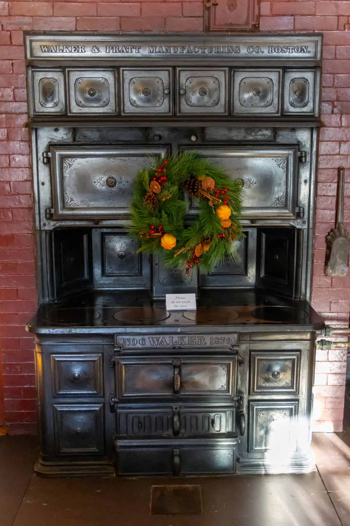 Antique cast iron stove with a decorative wreath hanging on its front, set against a brick wall. The stove is labeled "Walker & Pratt Manufacturing Co. Boston.