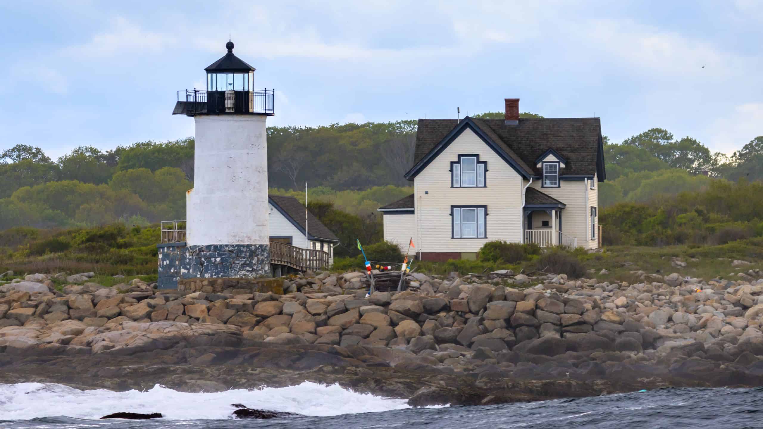 A white lighthouse and adjacent house stand on a rocky shoreline, with green trees in the background and waves crashing in the foreground—a picturesque example of lighthouses in Massachusetts.