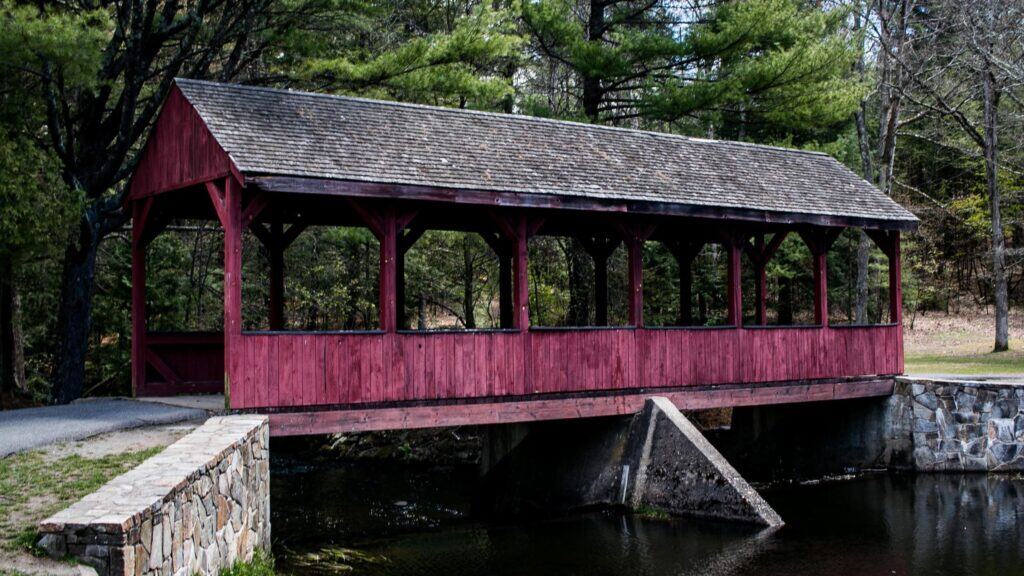 A red wooden covered bridge with a shingled roof extends over a creek, surrounded by trees and a stone wall on one side.
