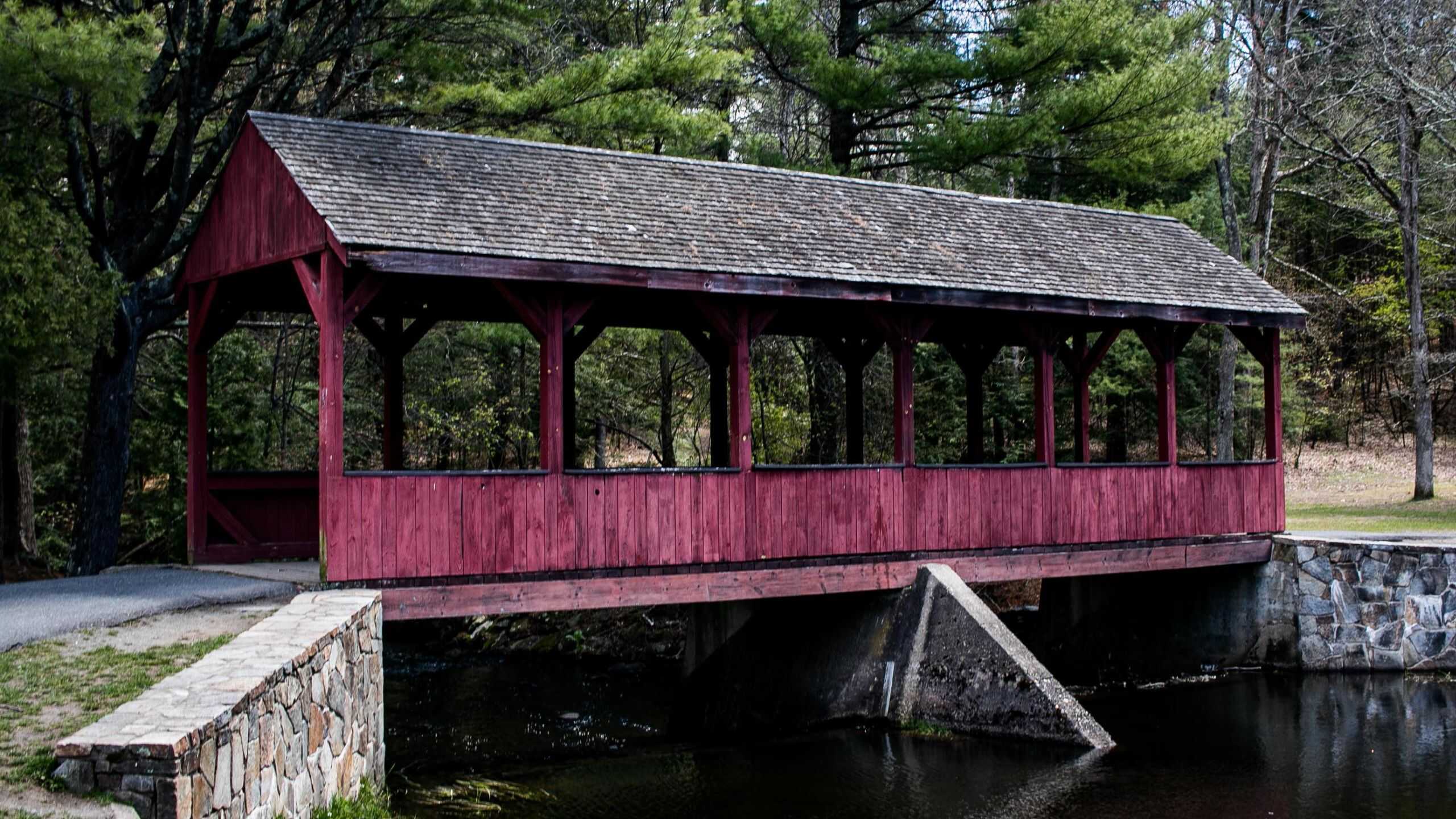 A red wooden covered bridge with a shingled roof extends over a creek, surrounded by trees and a stone wall on one side.