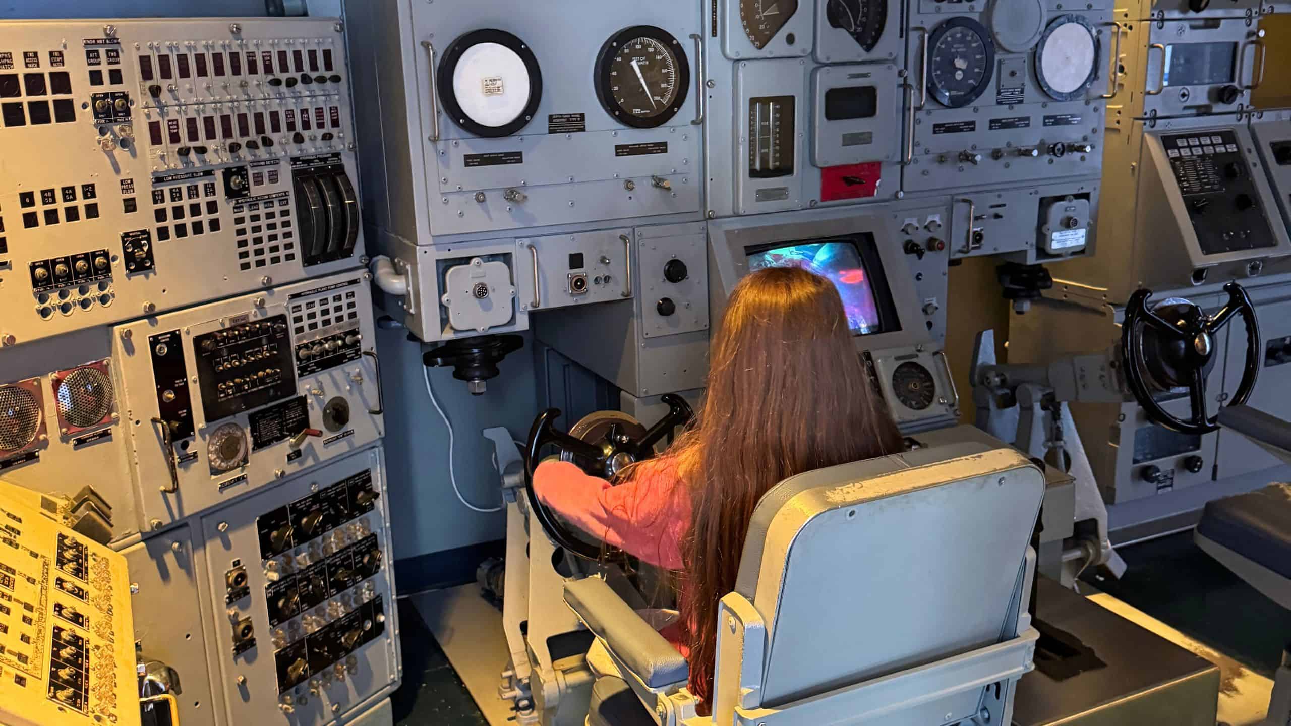A person with long brown hair sits at a control station with multiple dials, switches, and screens, operating the controls.