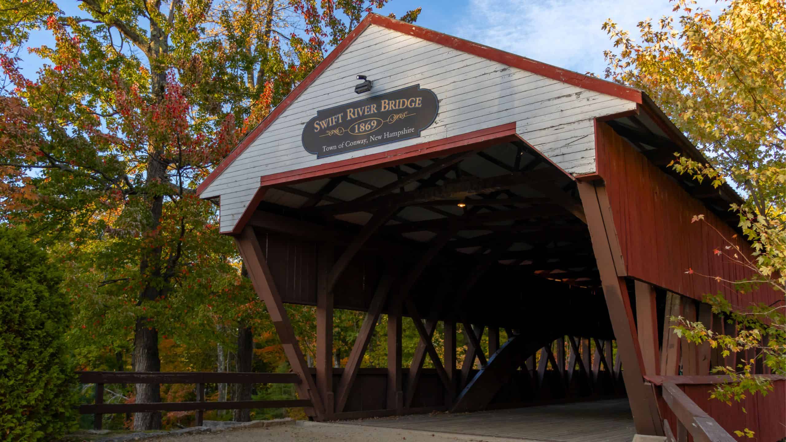 Covered wooden bridge with a “Saco River Bridge 1890” sign, surrounded by autumn trees in Conway, New Hampshire—a classic example of New England covered bridges.