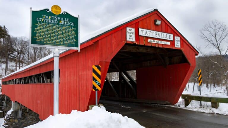 A red covered bridge with a sign reading "Taftsville Covered Bridge" is shown in a snowy landscape, with an informational plaque and traffic signs visible nearby.