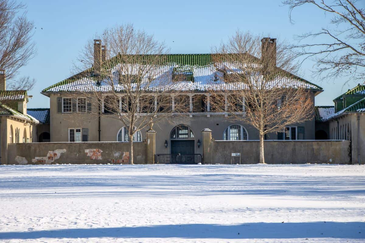 Large two-story house with green roof partially covered in snow, surrounded by a low wall and bare trees, with snow covering the ground.