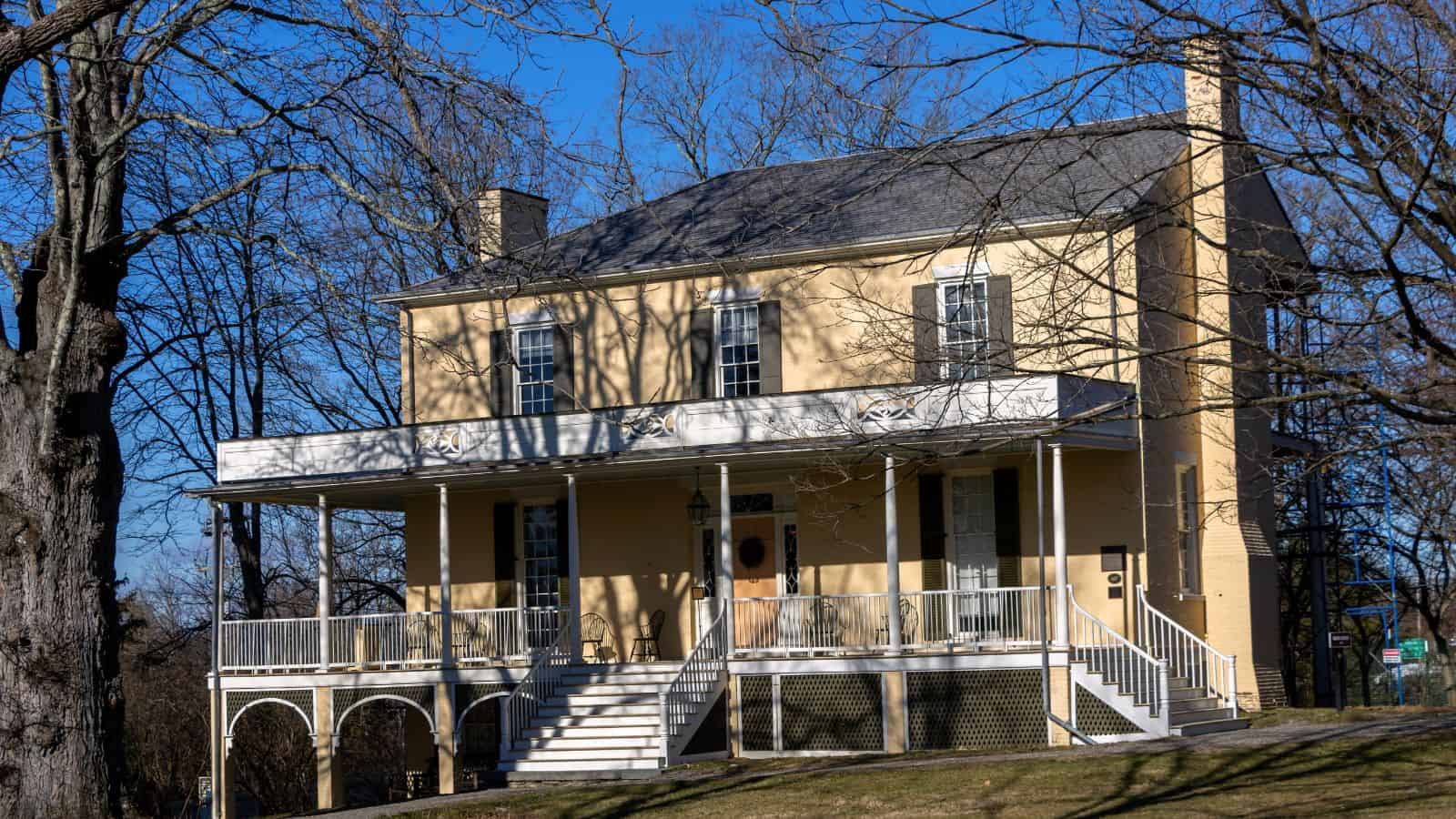 A two-story yellow brick house with a large front porch, white railings, and double stairs stands among leafless trees under a clear blue sky, evoking the charm of historic sites along the Hudson River.