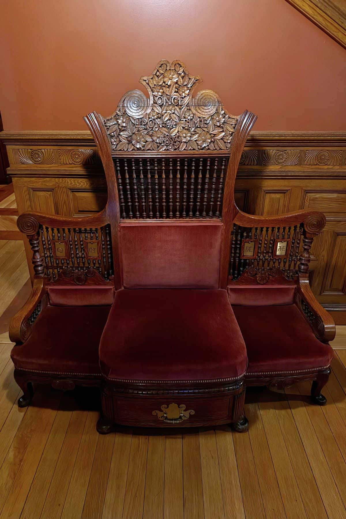 Ornate antique wooden three-seat bench with carved floral details, red velvet upholstery, and decorative armrests, placed on a hardwood floor against a wood-paneled wall.