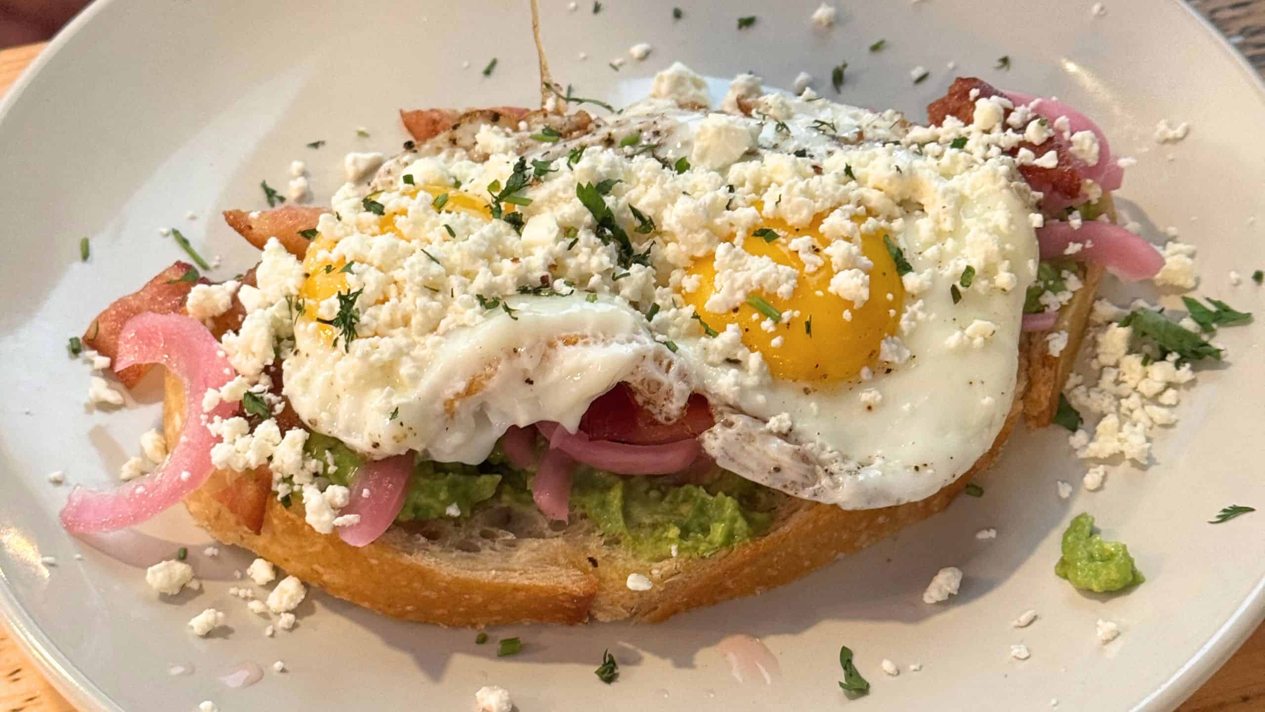 A slice of toasted bread topped with mashed avocado, bacon, pickled onions, two sunny-side-up eggs, crumbled cheese, and chopped herbs on a white plate.