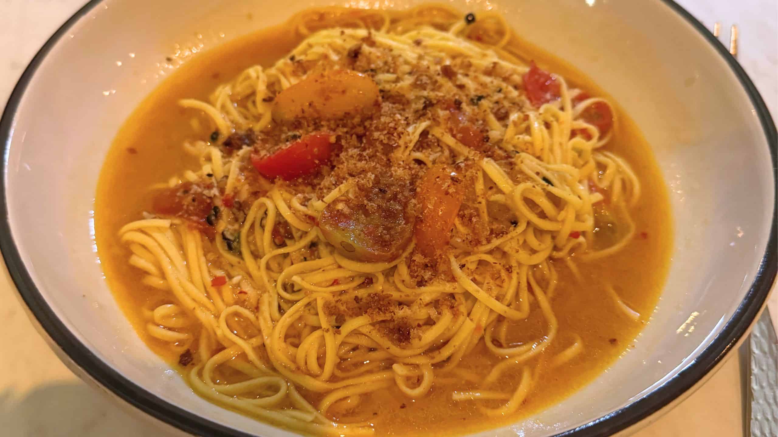 A bowl of noodle soup with yellow broth, garnished with chopped tomatoes and a sprinkle of brown crumbs.