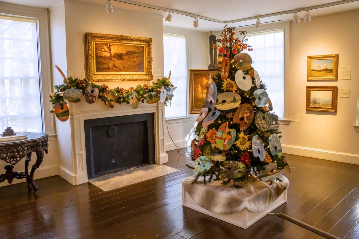 A decorated Christmas tree with artist palettes stands in an art gallery room with paintings on the walls and a mantel adorned with festive garlands.
