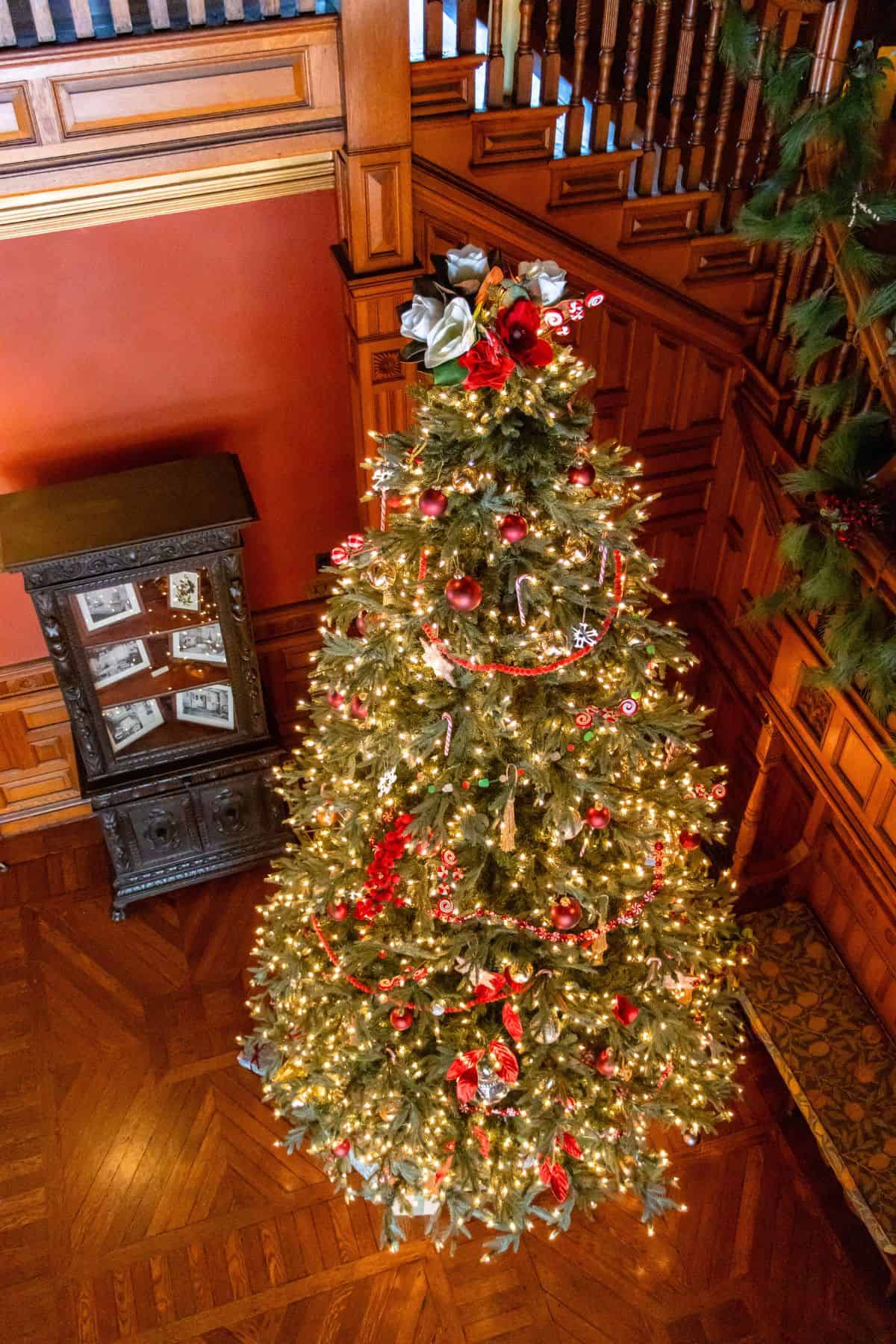 A decorated Christmas tree with lights, red ribbons, and ornaments stands in a wood-paneled room near a cabinet and staircase.