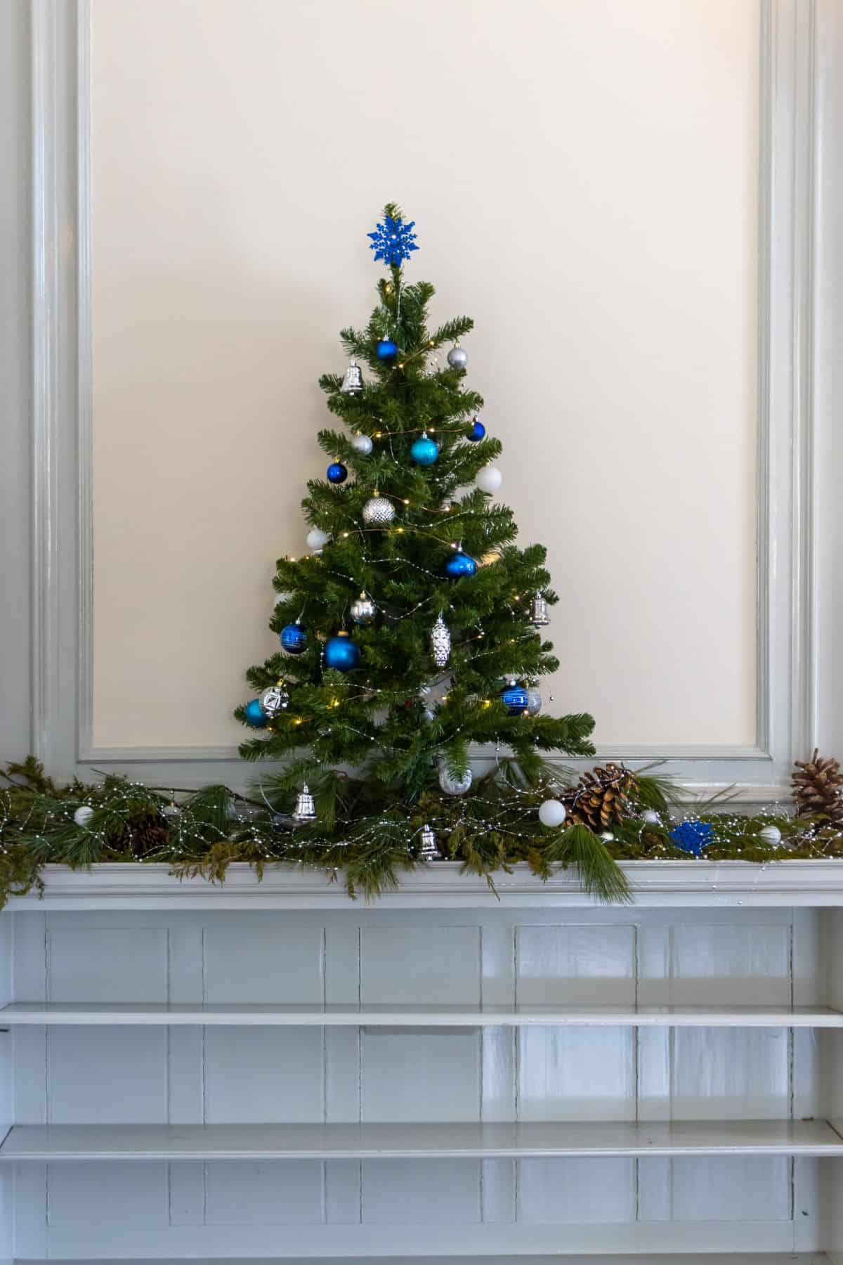 A small artificial Christmas tree decorated with blue and silver ornaments stands on a mantel, surrounded by garland and pinecones.