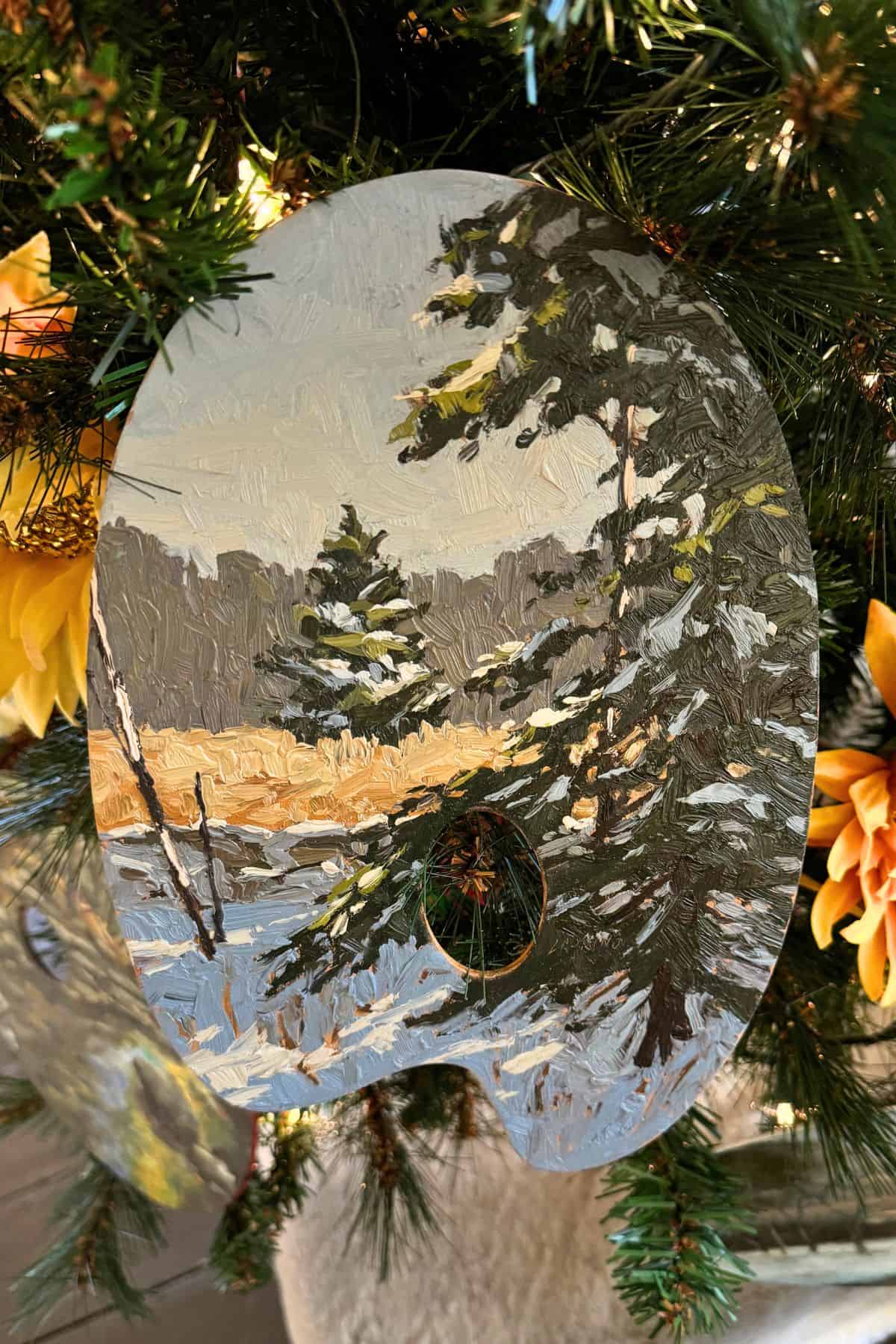 A paint palette shaped ornament with a winter landscape scene hangs on a decorated Christmas tree with green branches and yellow flowers.