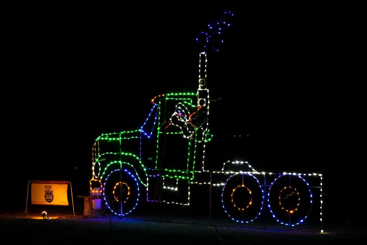 Outline of a semi-truck made with colorful holiday lights, featuring a Santa figure in the cab, displayed at night with a small illuminated sign nearby.