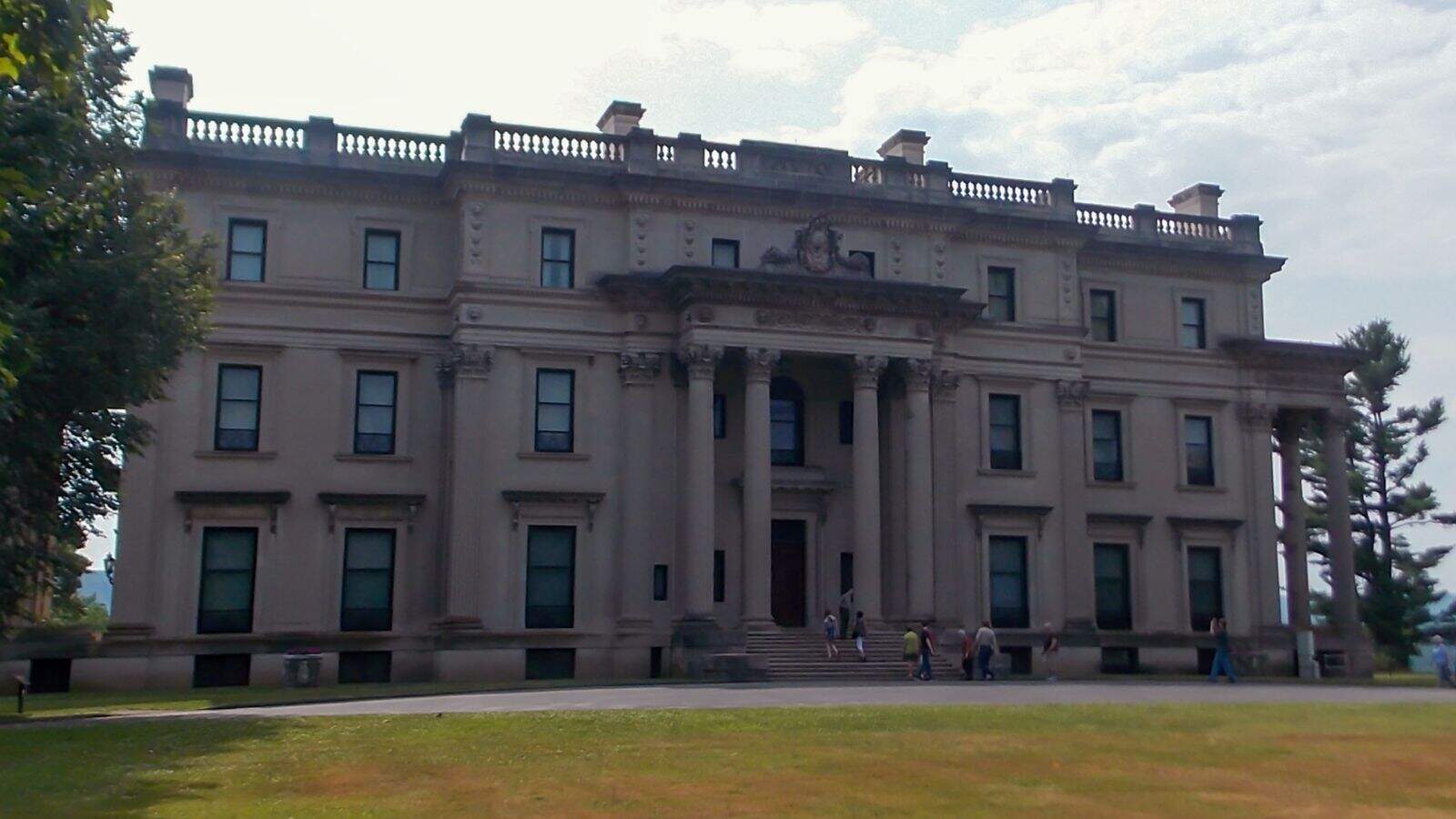 A large, three-story neoclassical mansion with columns at the entrance, several windows, and people walking in front on a sunny day.